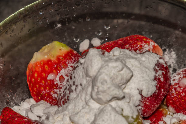 Making of young strawberries with sugar on black table in white tile kitchen