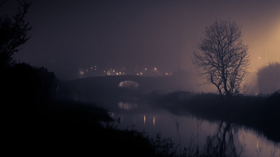 A dark foggy evening at the Royal Canal, Dublin, with eerie spooky scary tone, with distant bridge and tree illuminated by distant light