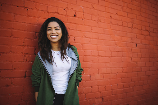 Natural light portrait of a beautiful, fashionable mixed race Latina, standing against the colored brick wall in Santa Monica, Los Angeles, California. She is just chilling, waiting for her friends, enjoying the nice summertime weather in California.