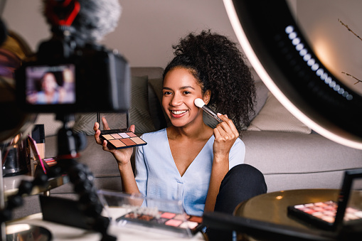 Smiling beauty blogger sitting in front of dslr camera with skin palette and brush in hands