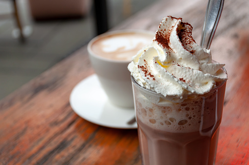 Tall glass of hot chocolate with whipped cream and spoon and cup of cappuccino on saucer in the background