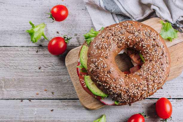 bagels com creme, abacate, tomates, rabanete e folhas verdes no fundo de madeira da placa e da tabela. comida saudável para o café da manhã. conceito equilibrado delicioso do alimento. vista superior e espaço da cópia - bun sandwich vegetable cream cheese - fotografias e filmes do acervo