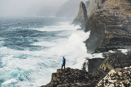 Young Caucasian man in blue raincoat looking at scenic  view of  the sea in Faroe Islands