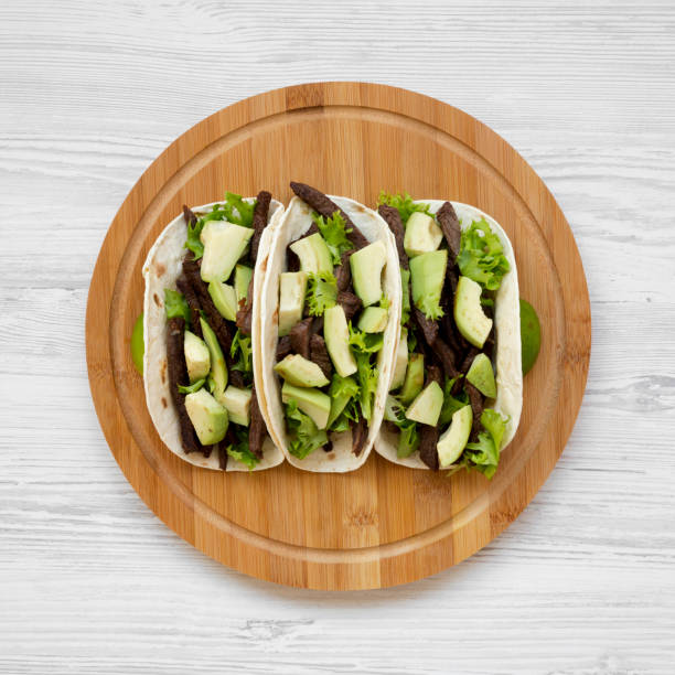 Corn tortillas with grilled beef, avocado, lime on round bamboo board, top view. Flat lay, from above, overhead.