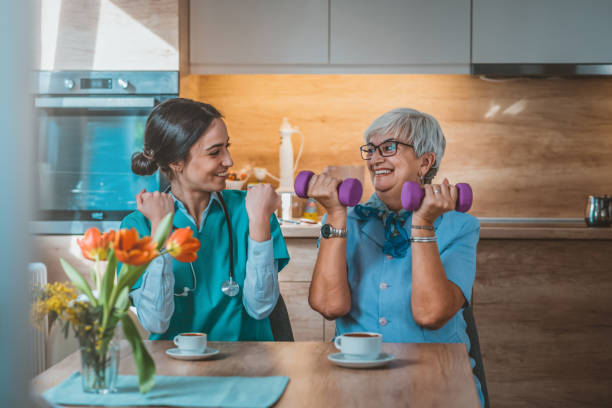 Nurse helping senior woman in lifting dumbbells Photo of Nurse helping senior woman in lifting dumbbell at retirement home during the day. Senior woman with dumbbells in rehab with a physiotherapist smiling. Physiotherapist helps elderly woman with healthy dumbbell training in rehab or home in the kitchen while sitting at table. occupational therapy photos stock pictures, royalty-free photos & images