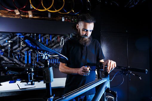Theme sale and repair of bicycles. Young and stylish with a beard and long hair, a Caucasian man uses a tool to set up and repair a bike in a store. Business owner at work.