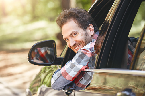Happy male driver in checkered shirt smiling while sitting in a car with open front window and looking back