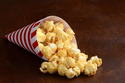 caramel popcorn in a red and white paper cone spilling on a wood table