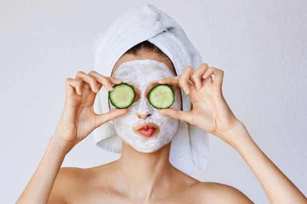 hermosa mujer joven con máscara facial en su cara sosteniendo rebanadas de pepino fresco - health or beauty fotografías e imágenes de stock