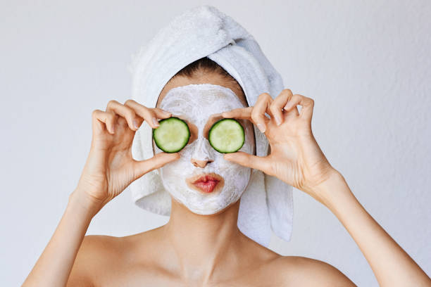 mooie jonge vrouw met gezichtsmasker op haar gezicht bedrijf plakjes verse komkommer - masker stockfoto's en -beelden