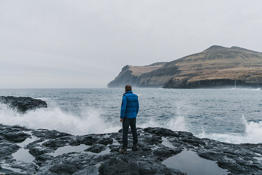 Young Caucasian man in blue raincoat  standing on the black sand beach and  looking at scenic  view of seaside in Faroe Islands