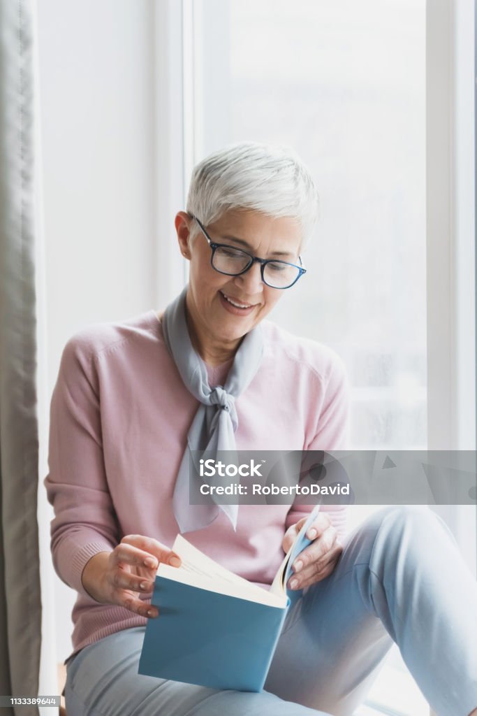 Mature woman reading book Attractive mature woman reading book in her bright modern cosy home, Enjoying free time and reading book concept Reading Stock Photo