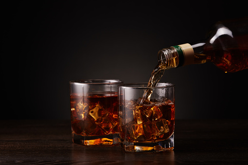 Pouring whiskey from a bottle into glass with ice standing on a dark table on a black background