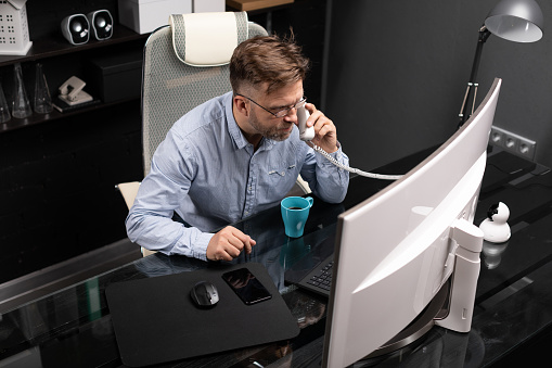 representative man with facial hair in gray shirt and jeans works in modern office at computer. businessman answers call on landline. Portrait of entrepreneur at work in office