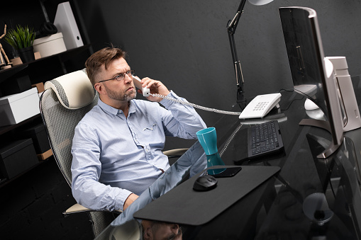 representative man with facial hair in gray shirt and jeans works in modern office at computer. businessman answers call on landline. Portrait of entrepreneur at work in office