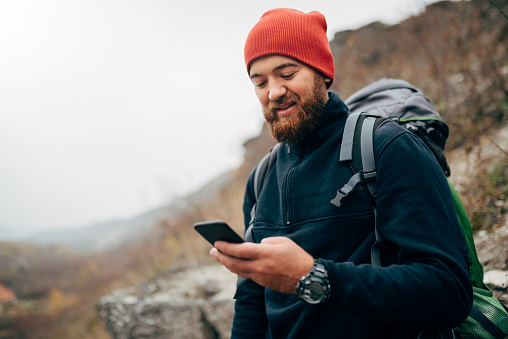 Young bearded man smiling and sending messages for his family from his cellphone, during hiking in mountains. Traveler bearded man in red hat using mobile phone application. Travel and lifestyle