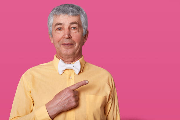 istock White haired mature man points aside at upper right corner, dresses yellow shirt with bow tie, isolated over pink background. Elderly male model poses in studio. People and measure concept. 1133442958