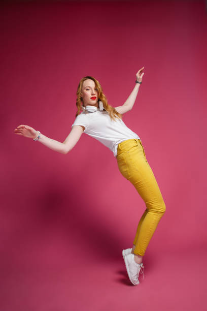 Cheerful blonde woman in white headphones, white T-shirt and trendy yellow jeans dancing on pink background stock photo