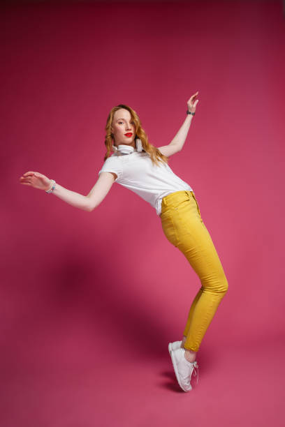 cheerful blonde woman in white headphones, white t-shirt and trendy yellow jeans dancing on pink background - shirt women pink jeans imagens e fotografias de stock