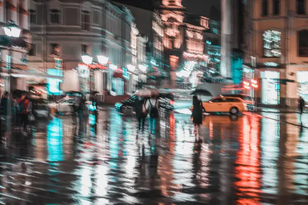 City street in rainy night, Abstract bright blurred background with unidentified people. Vivid illumination and reflection in wet pavement of shop windows and street lamps. Active lifestyle, leisure