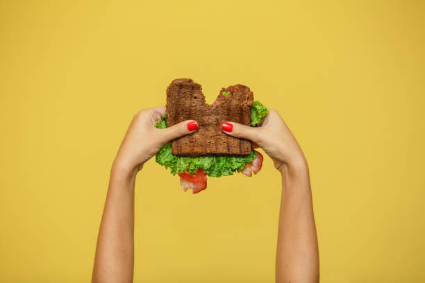 女性の手は黄色の背景にかまれたサンドイッチを保持します。サンドイッチプロモーションのコンセプト。 - appetizer lunch freshness vegetable ストックフォトと画像