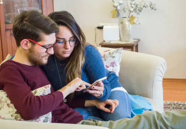 Lovely young couple using smartphone sitting on sofa in living room