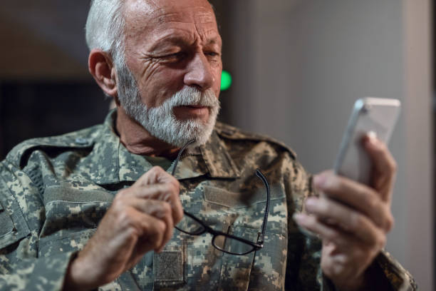 istock Smiling mature veteran in military uniform reading text message on mobile phone. 1133452384