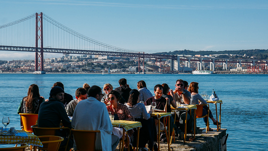 Lisbon, Portugal - March 2nd, 2019: People relax in outdoor restaurant terrace overlooking the iconic 25 April bridge in Lisbon, Portugal