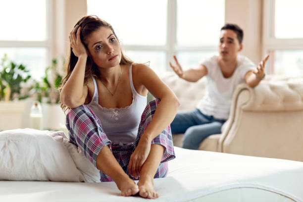 Disappointed woman thinking while sitting on bed and ignoring her boyfriend. Young sad woman with smudged make-up feeling heartbroken after the argument she had with her boyfriend. The man is in the background. woman mascara smudged stock pictures, royalty-free photos & images