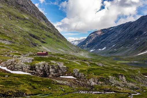 Majestic mountain view in Norway is captured on a sunny summer day. There is a small red house on the rocks surrounded by mountains.