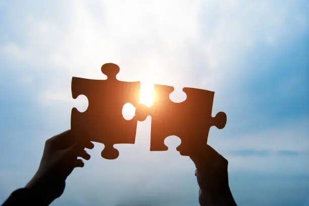 Human hands connecting two jigsaw puzzle pieces against cloudy sky.