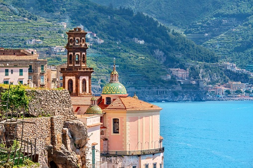 Dramatic church architecture on a steep cliff overlooking a vibrant blue sea, with green hills in the background. The church walls are painted a peach color, has tiled domes, and the brown bell tower adds elegance to the scene.