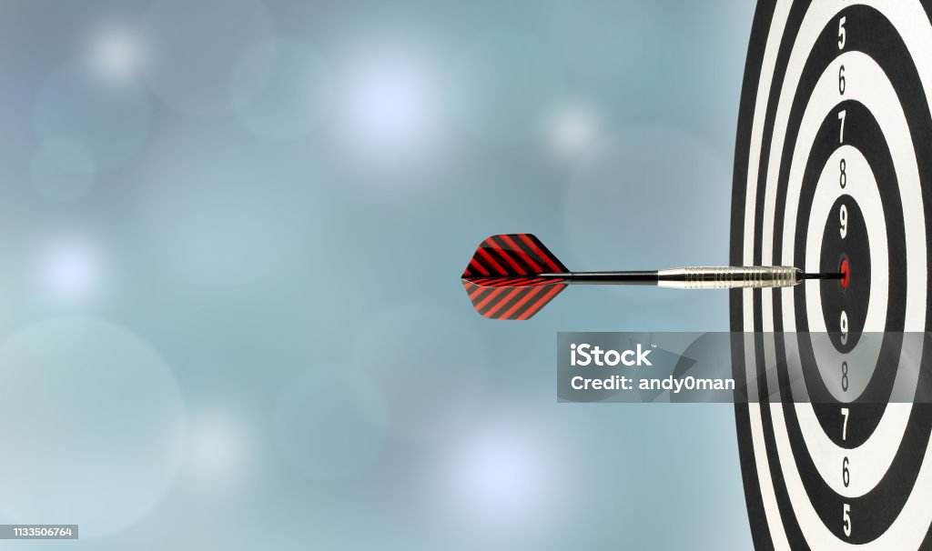 closeup silver metal dart arrow hitting red bulls eye target center of wooden dartboard with blurred blue lights bokeh copy space background perfection goal success, symbol of aim and achievement Sports Target Stock Photo
