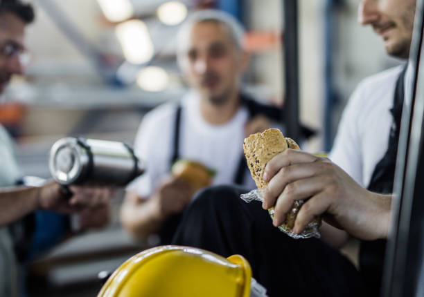 Close up of manual workers having a lunch break in a factory. Close up of worker eating sandwich while being on a lunch break with his coworkers in industrial building. construction lunch break stock pictures, royalty-free photos & images