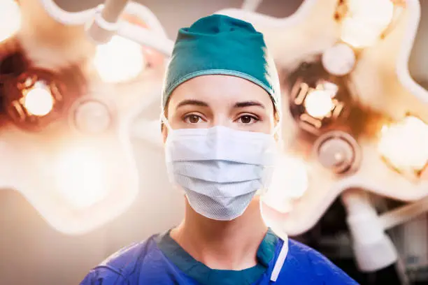 Portrait of female surgeon wearing surgical mask and cap. Skilled doctor is working in illuminated operating room. She is in hospital.