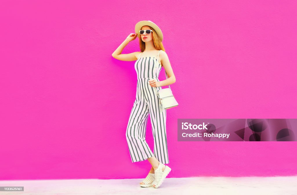 stylish woman model in summer round straw hat, white striped jumpsuit posing on colorful pink wall background Jumpsuit Stock Photo