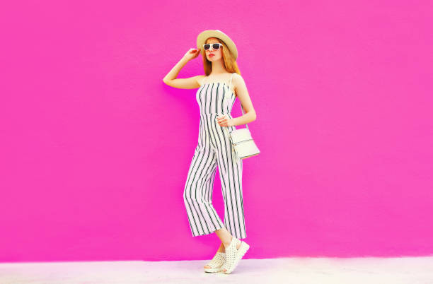 stylish woman model in summer round straw hat, white striped jumpsuit posing on colorful pink wall background stylish woman model in summer round straw hat, white striped jumpsuit posing on colorful pink wall background jumpsuit stock pictures, royalty-free photos & images