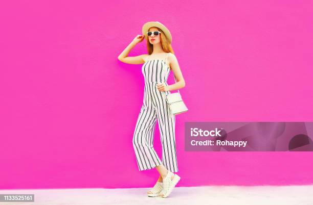 Stylish Woman Model In Summer Round Straw Hat White Striped Jumpsuit Posing On Colorful Pink Wall Background Stock Photo - Download Image Now