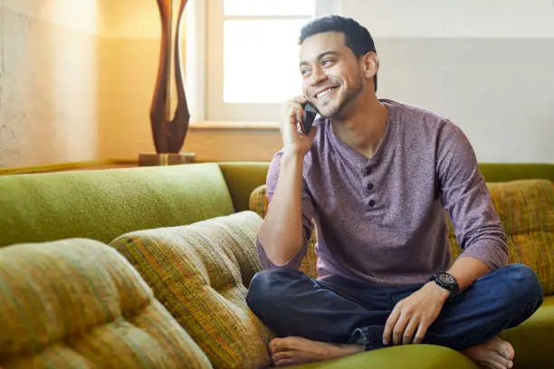 Smiling young man answering smart phone on couch. Happy confident male is looking away at home. He is enjoying conversation in living room.