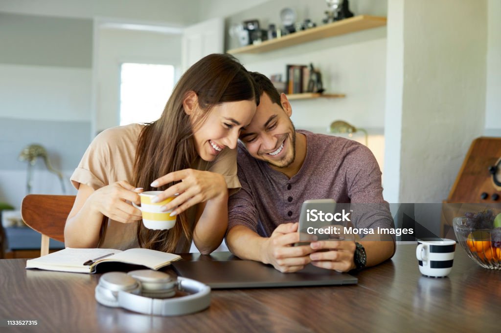 Cheerful couple using mobile phone in dining room Cheerful boyfriend showing mobile phone to girlfriend in dining room. Young woman is holding coffee cup. They are spending leisure time together at home. Couple - Relationship Stock Photo