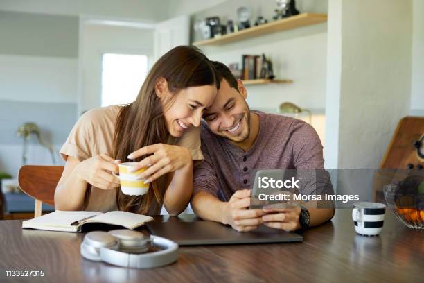 Cheerful Couple Using Mobile Phone In Dining Room Stock Photo - Download Image Now - Couple - Relationship, Mobile Phone, Domestic Life