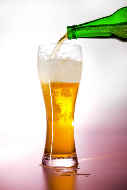 A bottle of green glass pouring beer into a beer glass. Background illumination with white light turning into light magenta. A bottle of green glass pouring beer into a beer glass. Background illumination with white light turning into light magenta. Close-up. кружка stock pictures, royalty-free photos & images