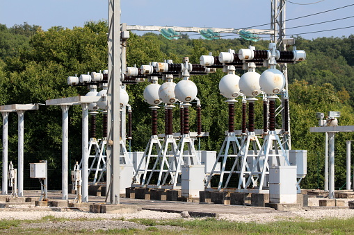 Dense array of electrical equipment at local power plant with strong metal support for dark brown ceramic and glass insulators connected with electrical wires surrounded with gravel and forest in background on warm sunny day