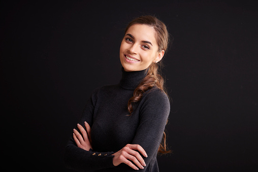 Close-up studio portrait shot of beautiful young woman wearing roll neck sweater and looking at camera while standing with arms crossed at dark background.