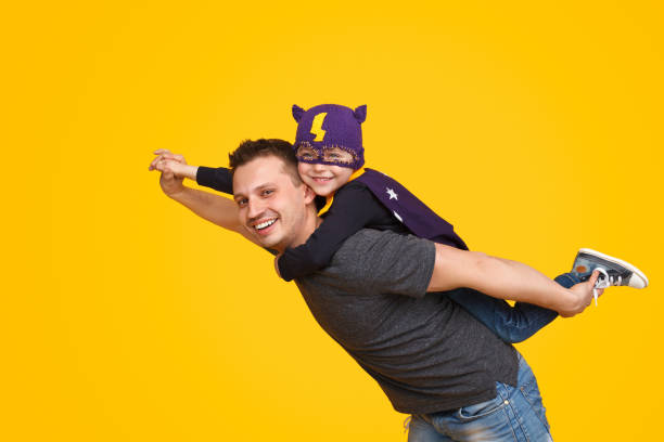 Father carrying little superhero on back Handsome man smiling and looking at camera while carrying cheerful boy in superhero mask on back on yellow background heroes photos stock pictures, royalty-free photos & images