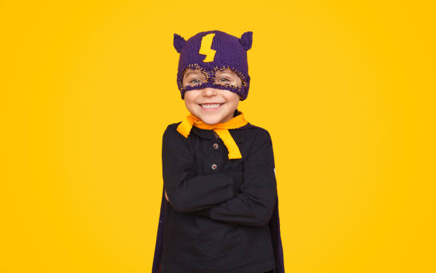 Adorable happy boy in knitted mask and cloak of hero smiling at camera on yellow background