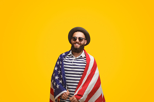 Trendy happy bearded man in hat covered with American flag smiling at camera on yellow backdrop