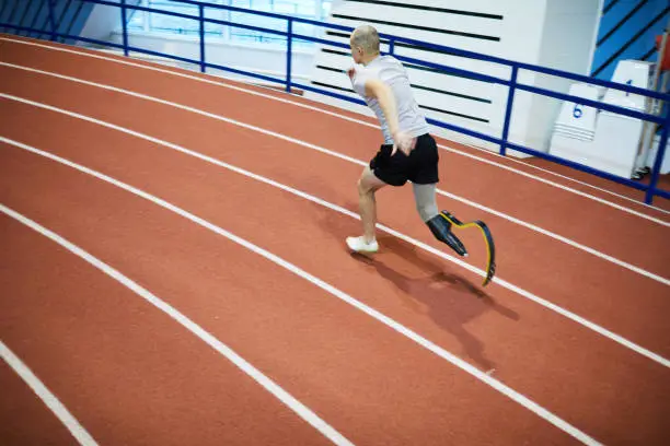 Young sportsman with prosthesis instead of right leg running down race track on stadium during competition