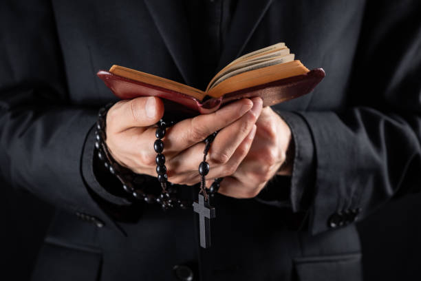 manos de un sacerdote cristiano vestido de negro sosteniendo un crucifijo y leyendo el libro del nuevo testamento. - catolicismo fotografías e imágenes de stock