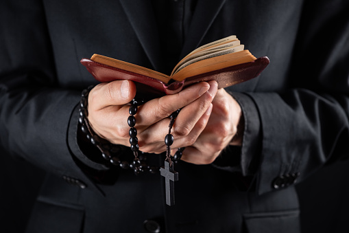 Hands of a christian priest dressed in black holding a crucifix and reading New Testament book.
